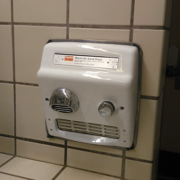 A slim white Dayton Warm Air Hand Dryer recessed into a tiled wall in a beige-coloured restroom