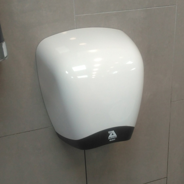 A bulbous white Airdri Quest Hand Dryer attached to a tiled wall in a grey restroom