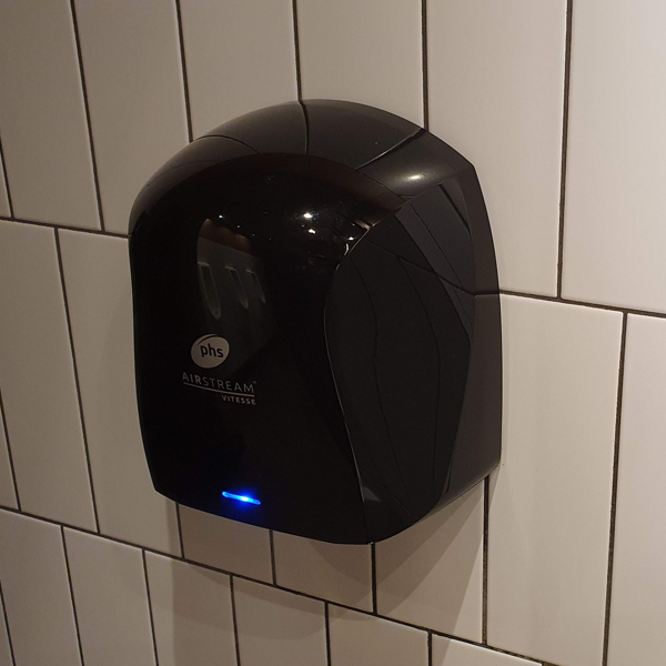 A sleek black Airstream Vitesse Hand Dryer attached to a tiled white wall in a modern restroom