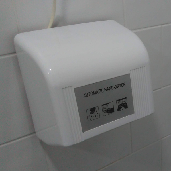 A basic white Automatic Hand-Dryer attached to a white tiled wall in a simple clean restroom