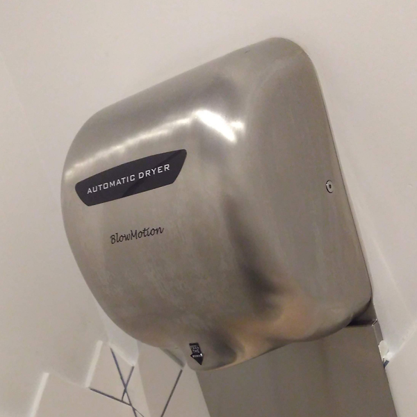 A thicc silver Blowmotion Hand Dryer attached to a plain white wall in a modern restroom