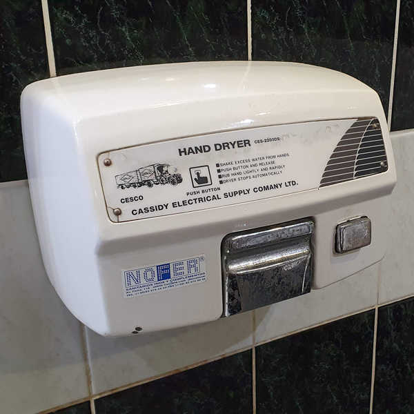 A chunky white CESCO Hand Dryer attached to a black and white tiled wall in a retro restroom