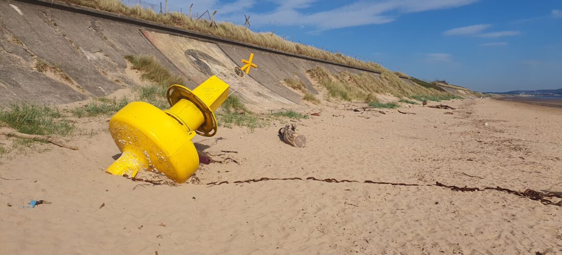 A large yellow buoy lies tilted on a sandy beach near a grassy embankment under a blue sky. The scene conveys a peaceful, abandoned atmosphere.